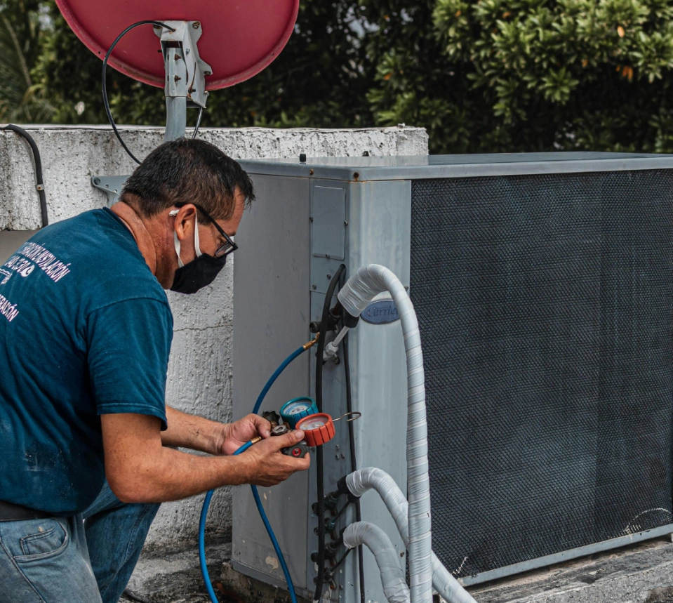 Man in a face mask using gauges to service an outdoor air conditioning unit on a rooftop.